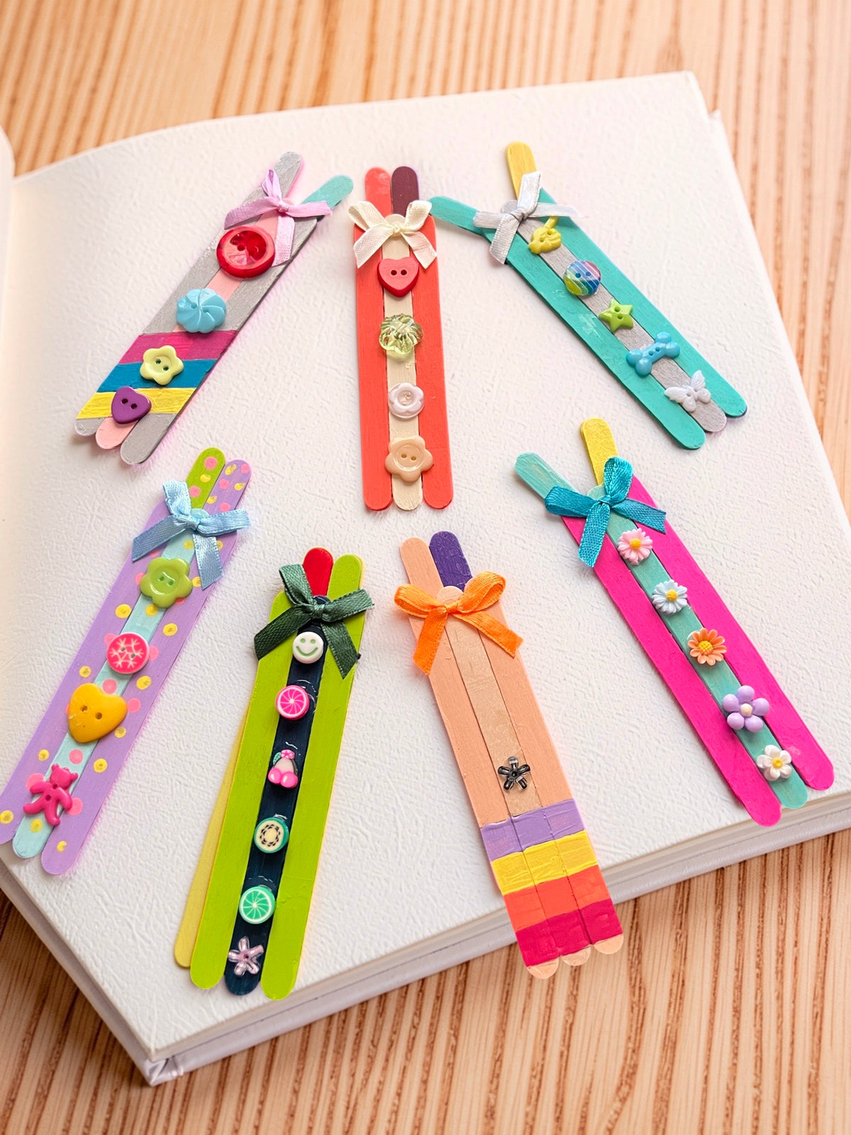 "Seven colorful handcrafted popsicle stick bookmarks with ribbon bows, buttons, and flower embellishments arranged on a white book"