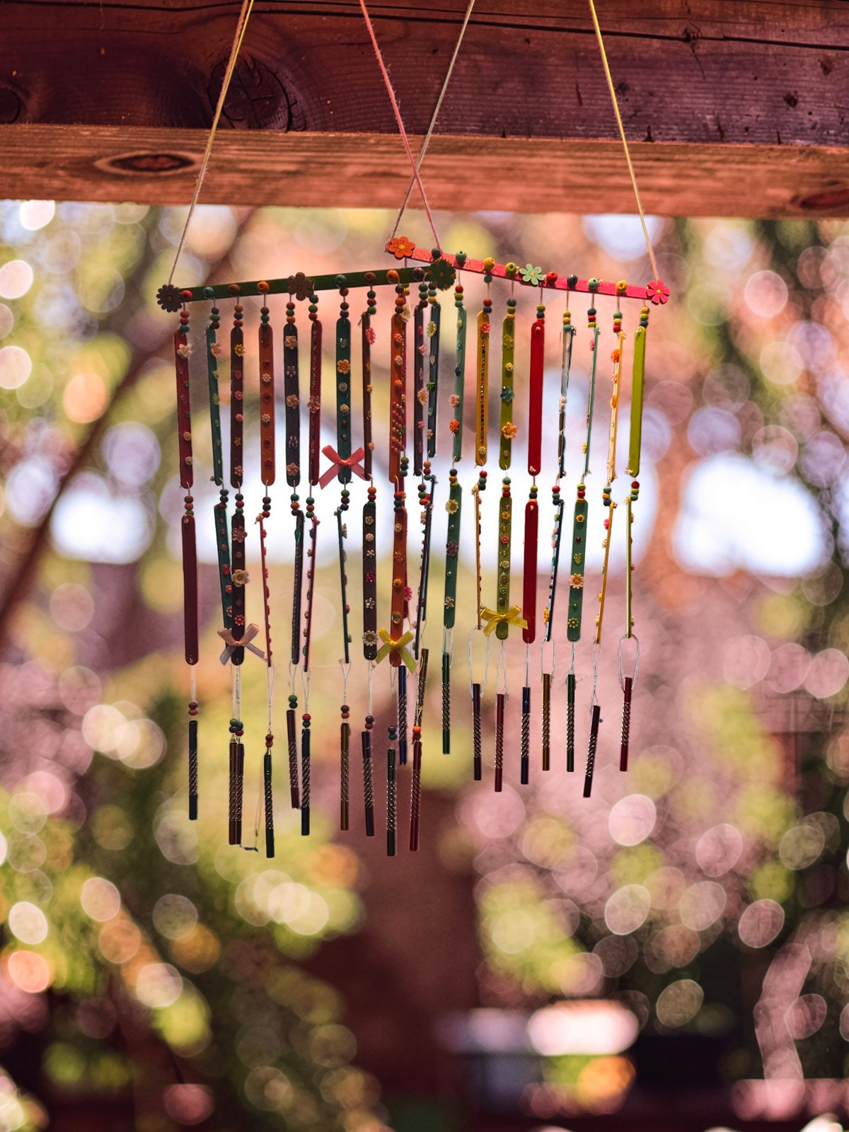Two sets of large wooden wind chimes decorated with colorful jewels, charms, and flowers, displayed against a soft-focus forest background.