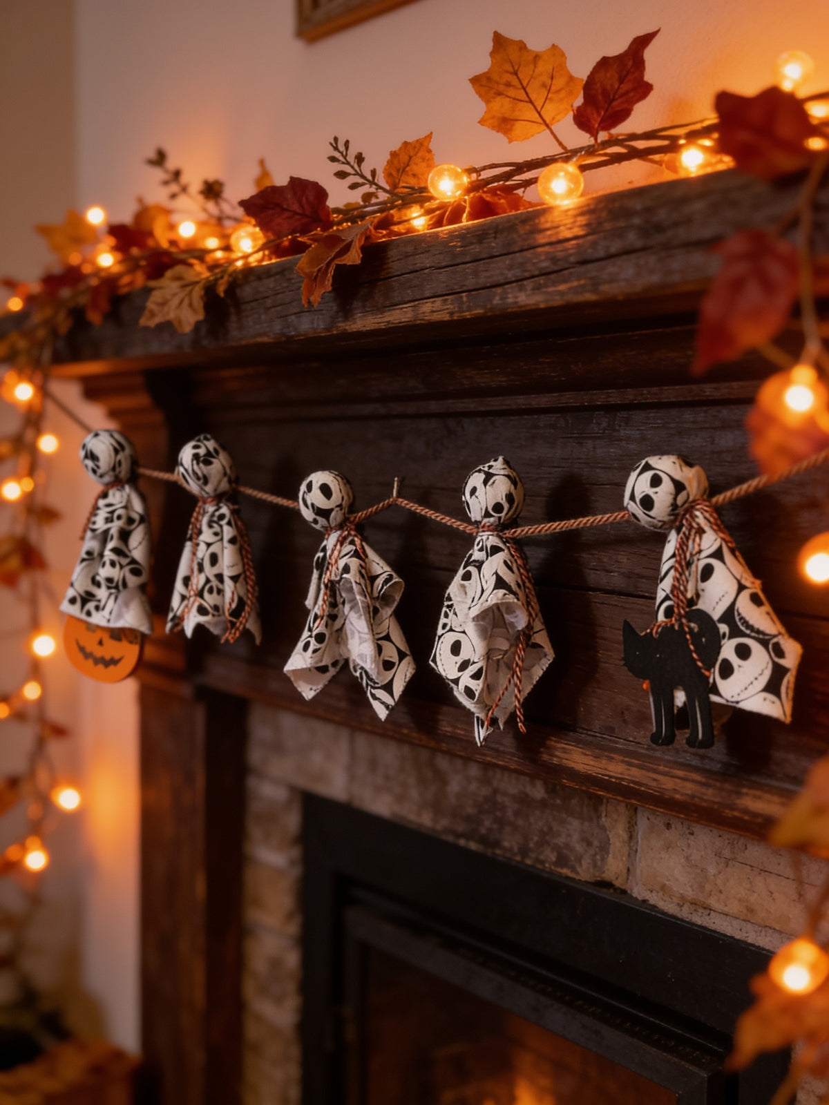 Five black-and-white fabric ghosts on twine with pumpkin and black cat accents, displayed across a mantel with string lights.