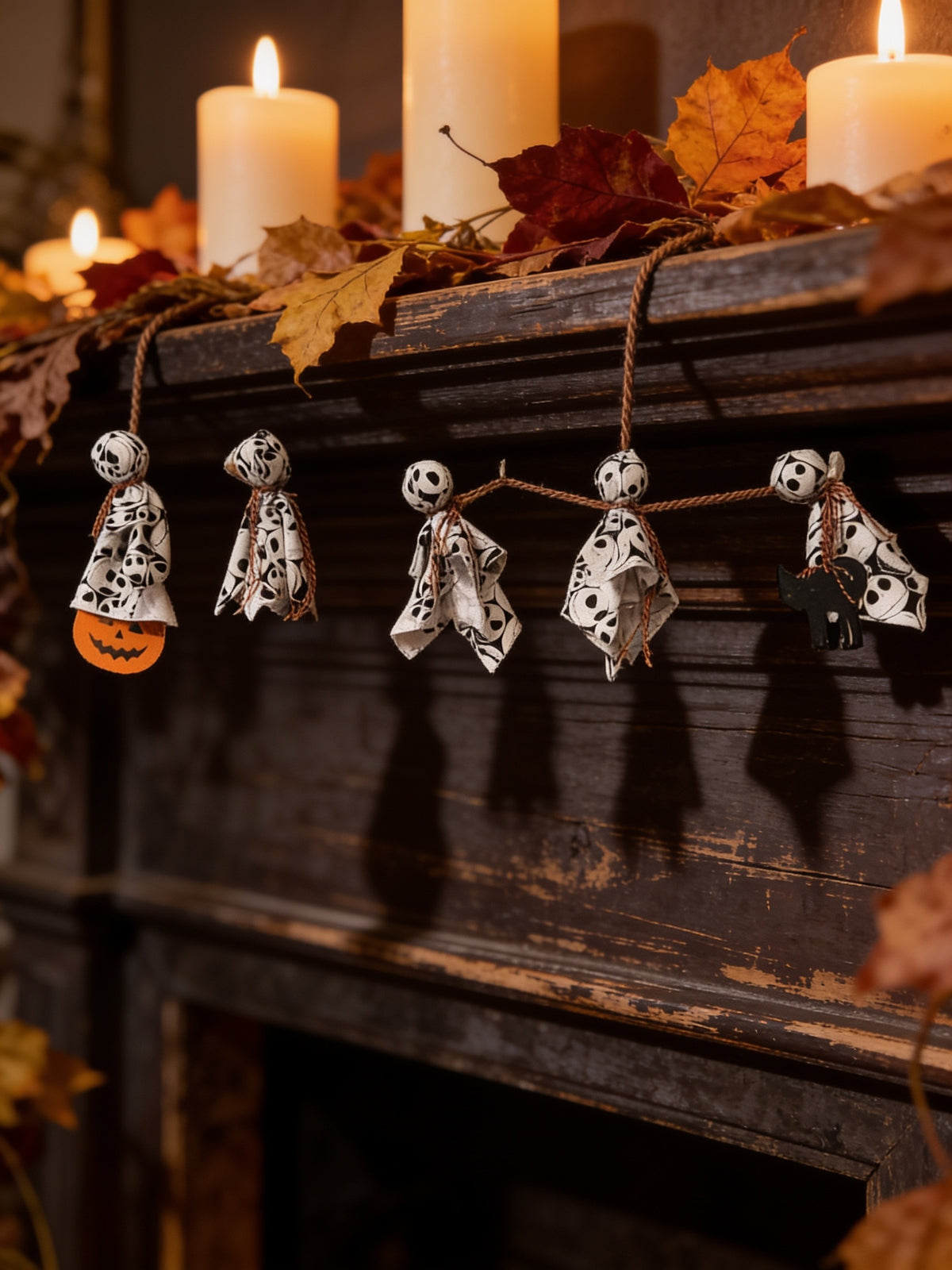 Halloween streamer with five patterned fabric ghosts, a pumpkin and black cat, hanging across a wooden mantel beneath candles.