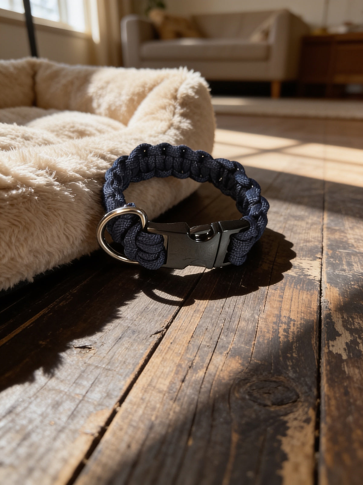 Small dark blue woven paracord dog collar with buckle and metal D-ring displayed beside a plush dog bed.