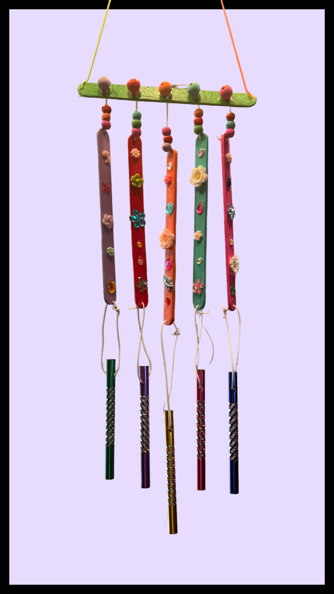 Small wooden wind chimes decorated with colorful jewels, charms, and flowers, displayed within a black frame against an off-white background.