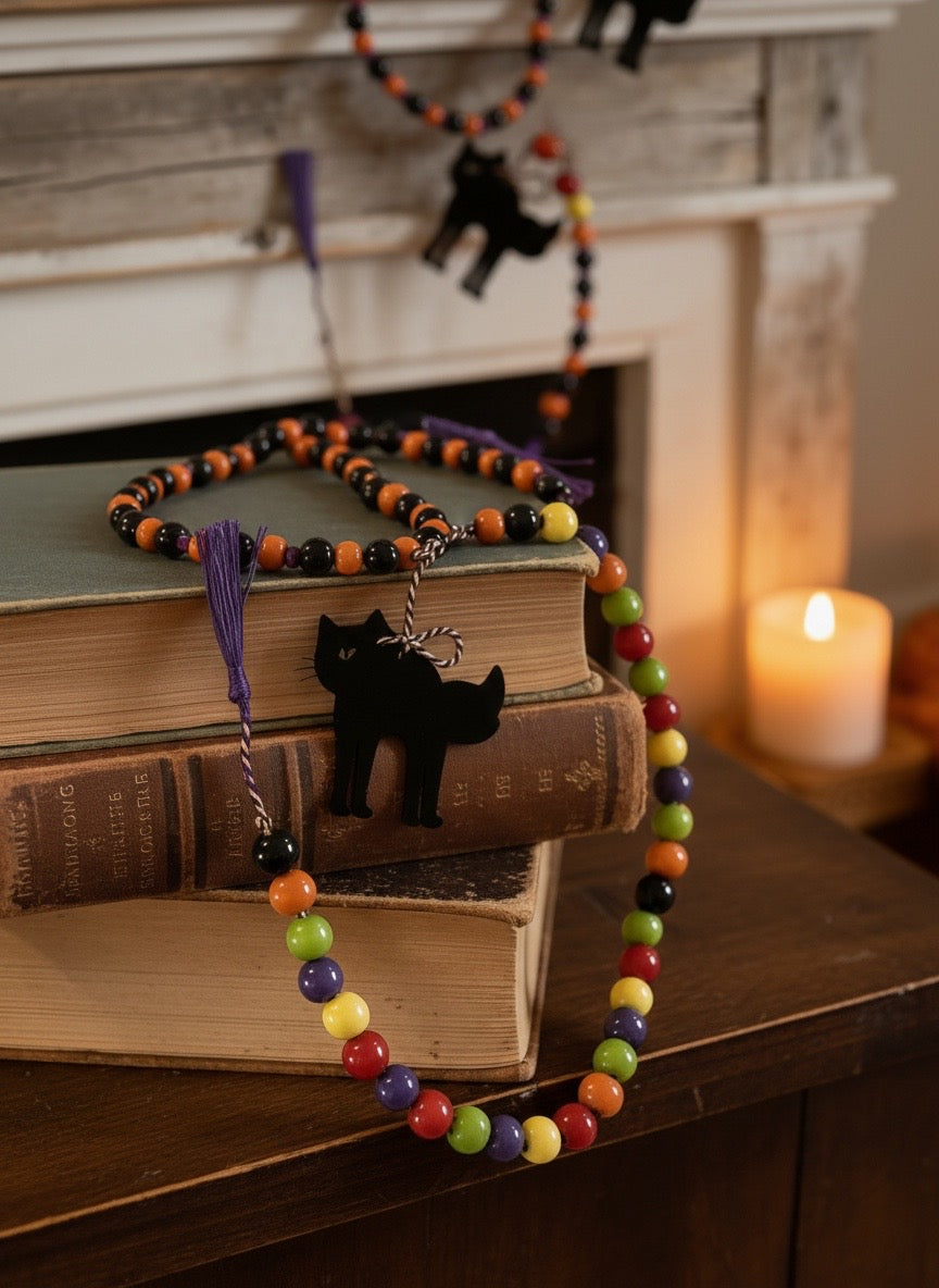 Handmade Halloween beaded garland with a black cat wood piece and purple tassel resting on a stack of old books, shown with a second garland above.


