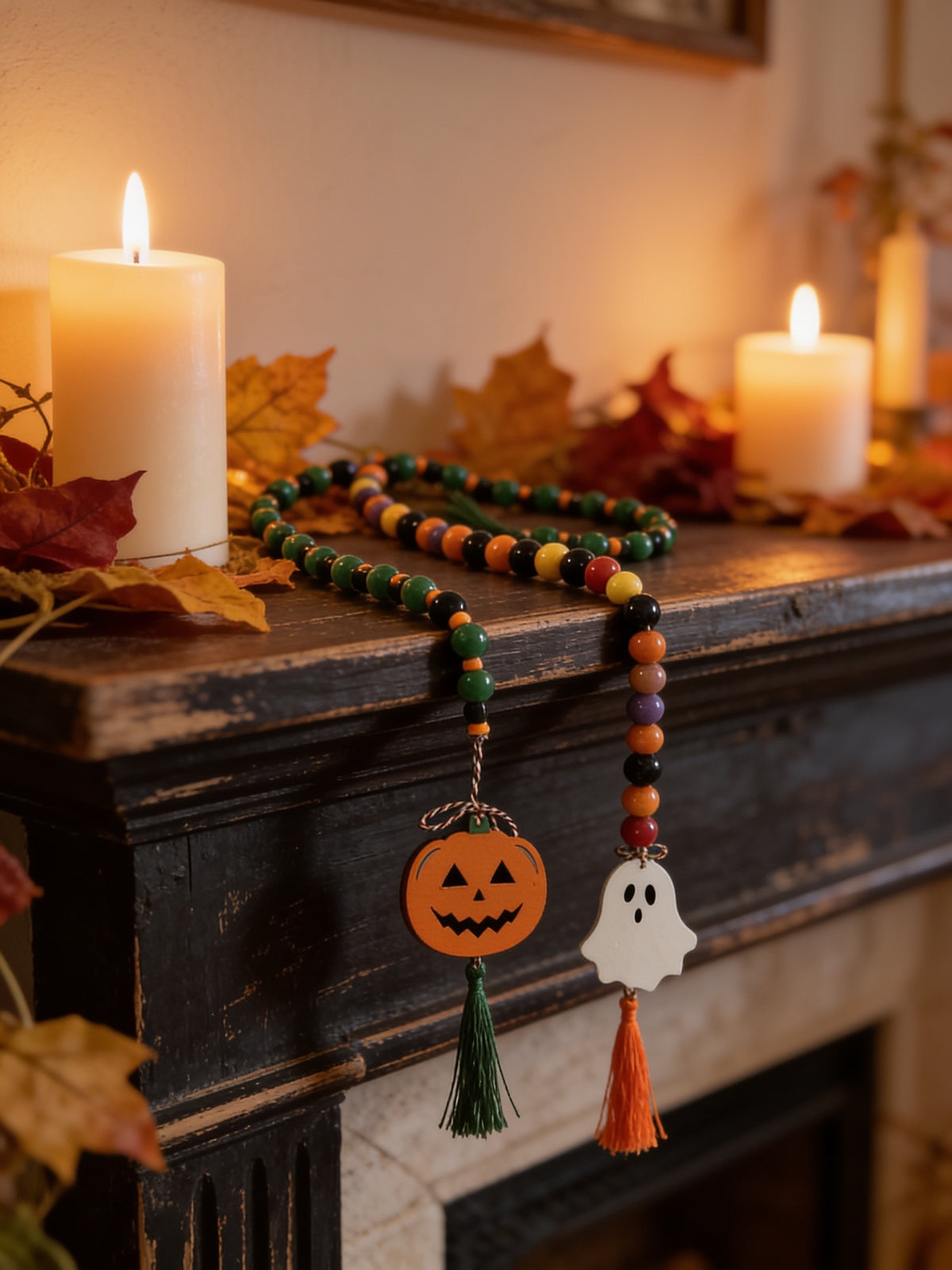 "Two Halloween beaded garlands on a dark mantel, one in green and black with an orange jack-o'-lantern and green tassel, one in black, red, orange and purple with a white ghost and orange tassel, beside lit candles and fall leaves."